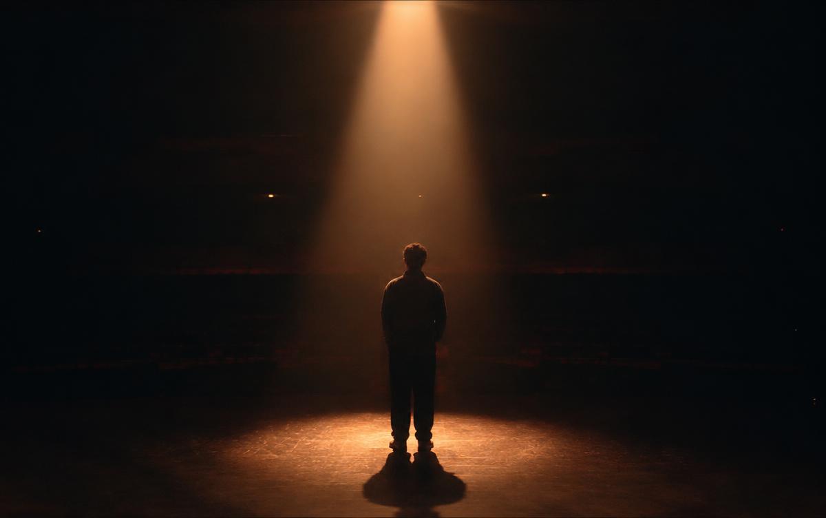 Cinematic still of a lone figure under a warm spotlight in a dark theatre